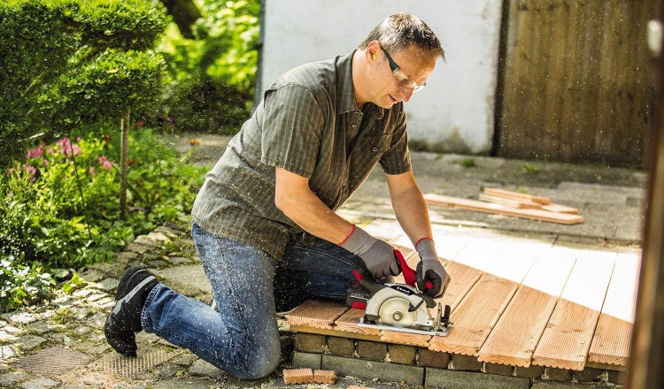 Handwerker mit Schutzausrüstung sägt Holzbretter für eine Terrasse mit einer Akku Handkreissäge im Freien.