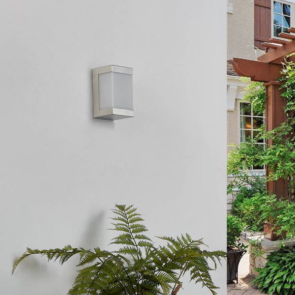 Moderne Außenwandleuchte aus Edelstahl mit weißem Schirm an einer Hauswand, im Hintergrund ein Garten mit einer hölzernen Pergola.