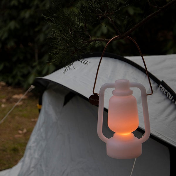 Leuchtende weiße LED-Campinglaterne mit Drahtbügel, hängend an einem Ast vor einem Zelt im Freien.