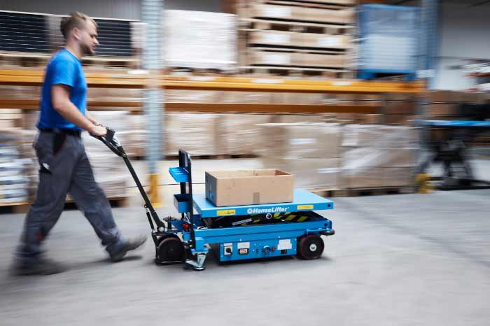 Ein Mann schiebt einen blauen HanseLifter Scherenhubwagen mit einem Karton durch ein Lager mit Hochregalen.