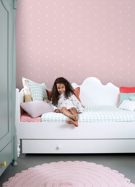 Kinderzimmer mit Bett, rosa Teppich und Mädchen vor einer mit Kreuzen verzierten Tapete