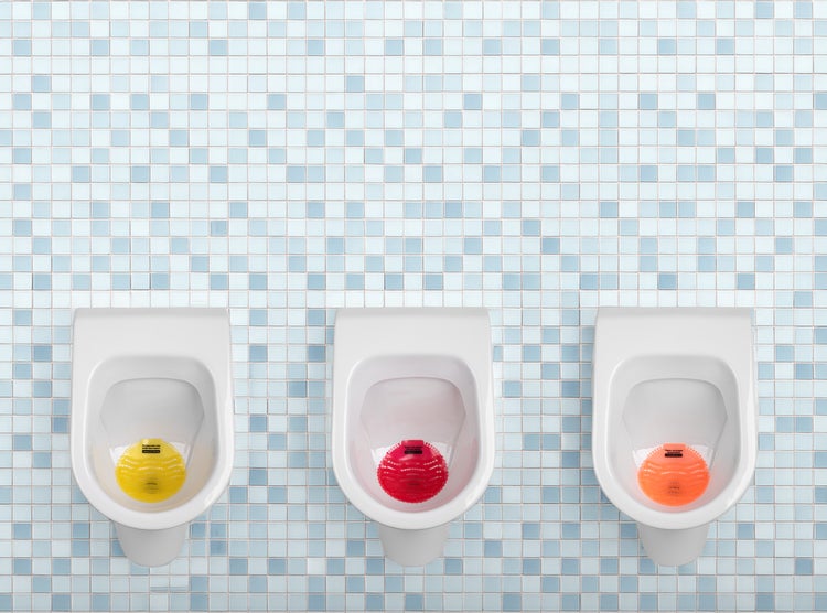 Drei Urinale in Reihe vor einer gefliesten Wand mit jeweils einem farbigen Urinalsieb zur Geruchshemmung und Spritzschutz.