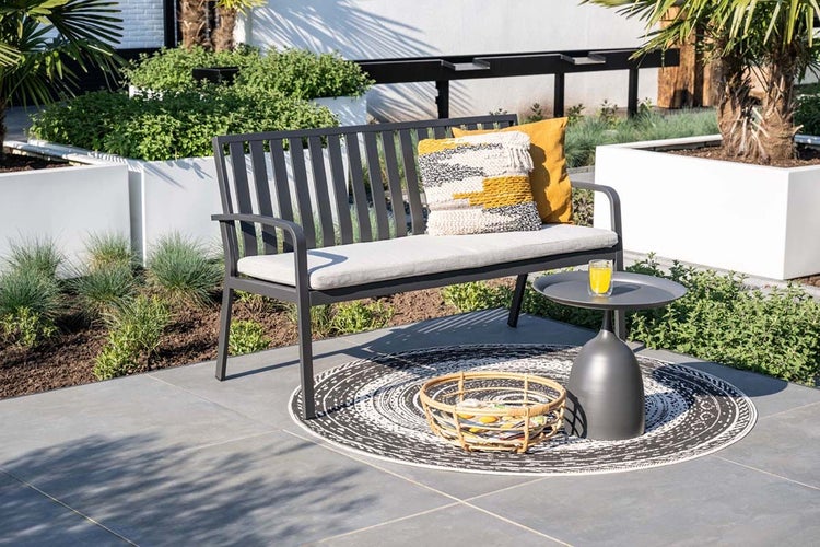 Moderne Gartenbank aus Metall mit grauer Sitzauflage und Zierkissen auf einer Terrasse, kleiner Beistelltisch und Teppich.