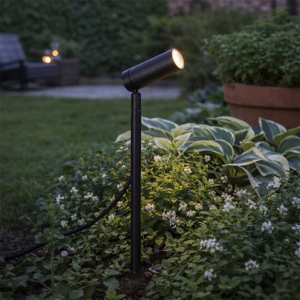 Schwarzer LED Gartenstrahler mit Erdspieß beleuchtet grüne Pflanzen und weiße Blumen in einem Garten bei Dämmerung.
