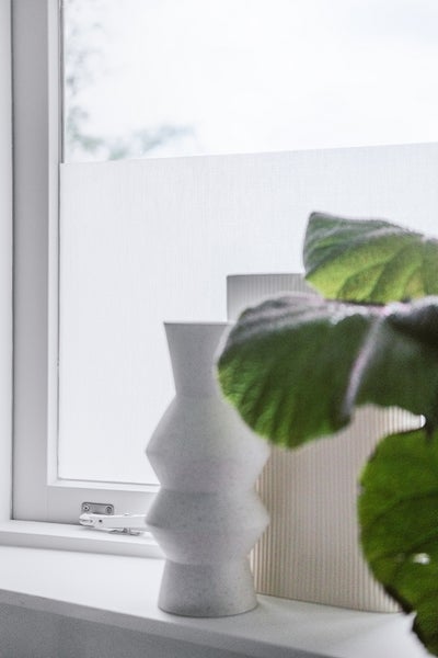 Helles Fenster mit Sichtschutzfolie auf der Scheibe, einer geometrischen Vase und einer Pflanze auf der Fensterbank.