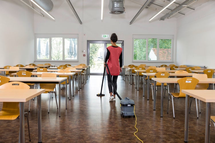 Reinigungskraft saugt den Boden in einem großen hellen Raum mit vielen Tischen und Stühlen mit einem professionellen Staubsauger.