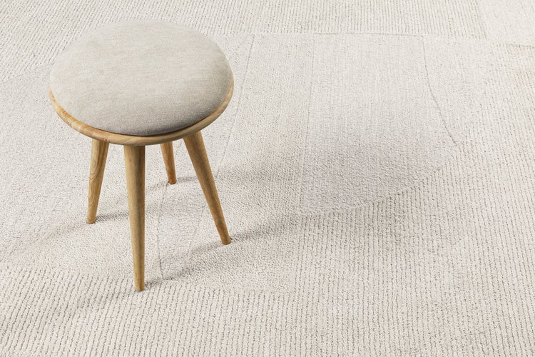 Runder Hocker mit Holzbeinen und gepolsterter Sitzfläche auf einem hellen, strukturierten Teppich mit geometrischem Muster.