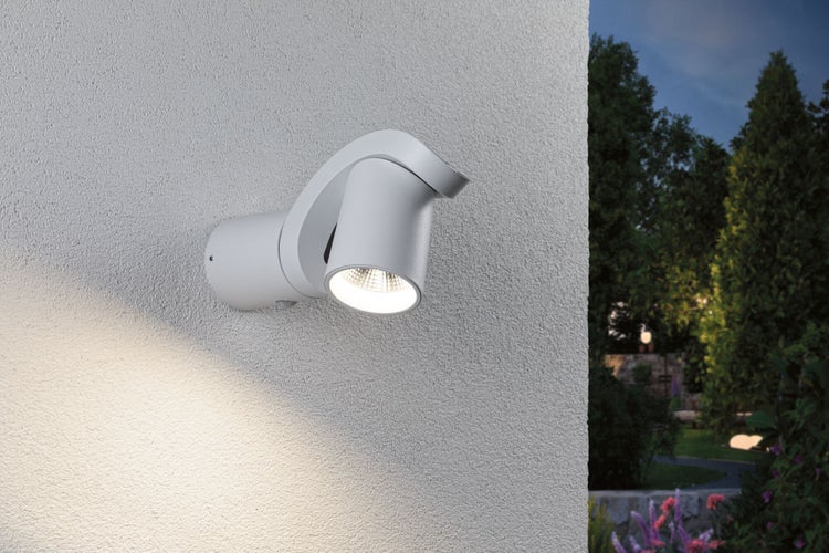 Moderner, verstellbarer LED-Außenstrahler in Weiß an einer Wand montiert, eingeschaltet bei Nacht vor einem Gartenhintergrund.