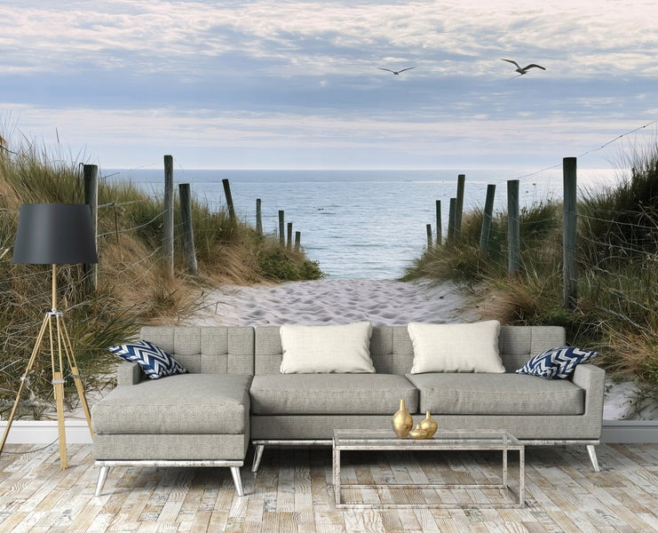 Fototapete mit Dünenpfad am Meer im Wohnzimmer mit grauem Sofa, Stehlampe und Couchtisch, modernes Wanddesign mit Strandmotiv.