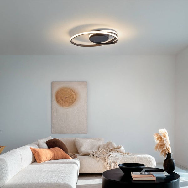 Modernes Wohnzimmer mit ringförmiger LED-Deckenleuchte, hellem Ecksofa, orangefarbenen Kissen und schwarzem Couchtisch in heller Atmosphäre.