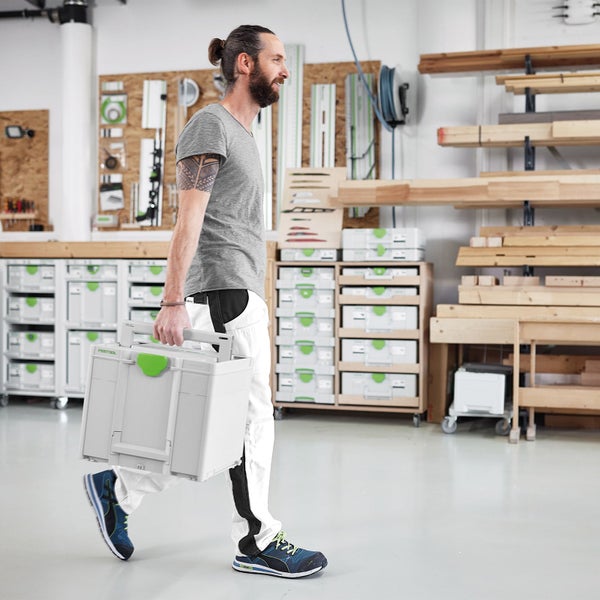 Ein Mann trägt einen großen grauen Werkzeugkoffer in einer organisierten Werkstatt mit zahlreichen Systainern und Holzvorräten.