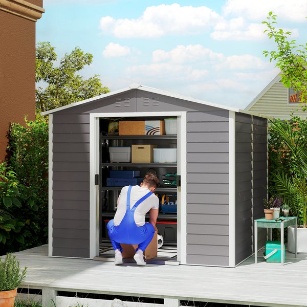 Modernes graues Gerätehaus aus Metall mit geöffneter Tür auf Holzterrasse. Ein Mann in blauer Latzhose räumt im Inneren Regale mit Werkzeug ein.
