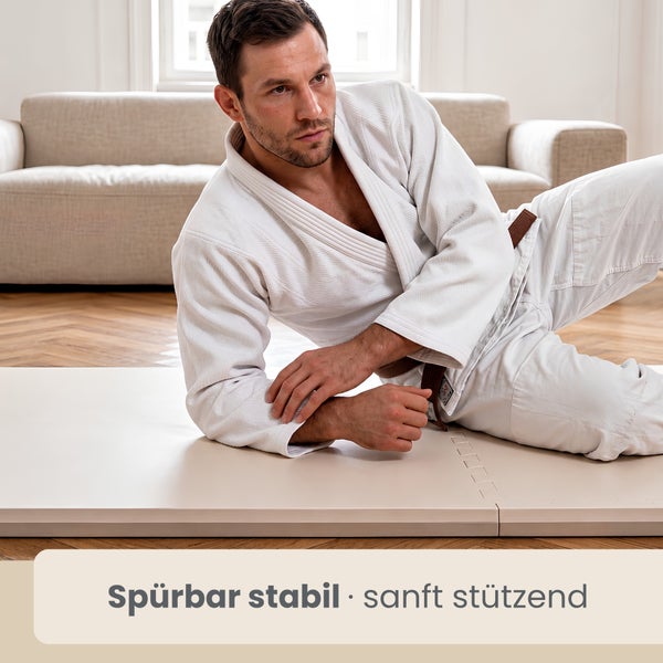 Ein Mann im Kampfsportanzug auf einer stabilen Bodenmatte in einem hellen Wohnzimmer mit Sofa.