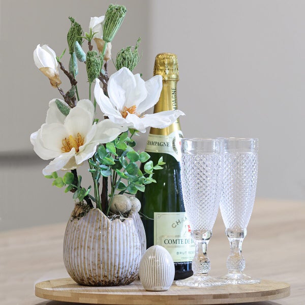Dekoratives Arrangement mit künstlichen Magnolien in einer Vase, Champagnerflasche, zwei Sektgläsern und Osterei auf einem runden Holztablett.