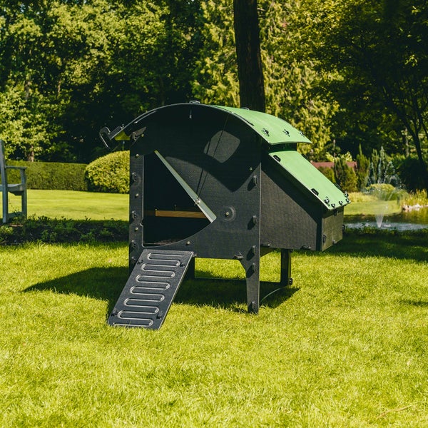 Hühnerstall mit grünem Bogendach und Rampe auf einer Rasenfläche in einem hellen Garten.