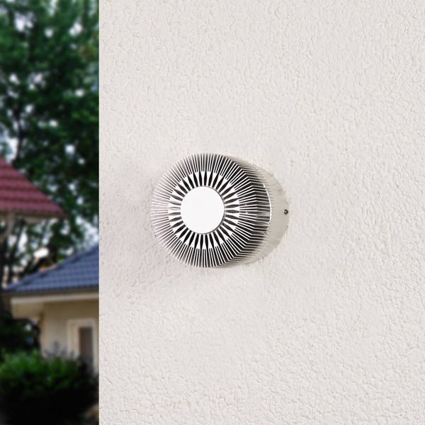Runde, silberne Außenwandleuchte aus Metall mit radialem Lamellendesign an einer strukturierten Fassade.