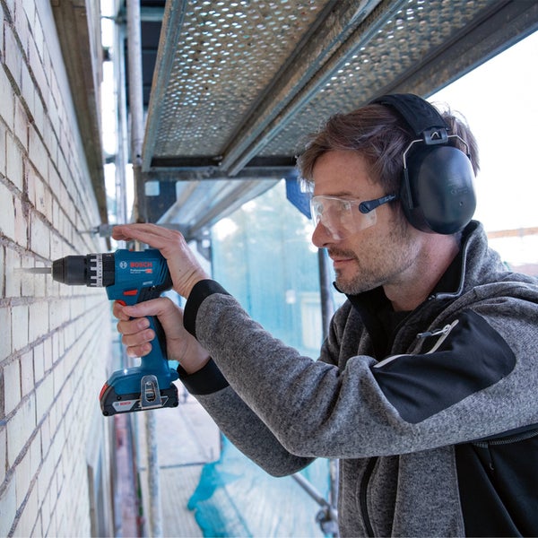 Ein Handwerker bohrt mit einer Bosch Akku-Bohrmaschine ein Loch in eine Ziegelwand. Er trägt dabei eine Schutzbrille und einen Gehörschutz.