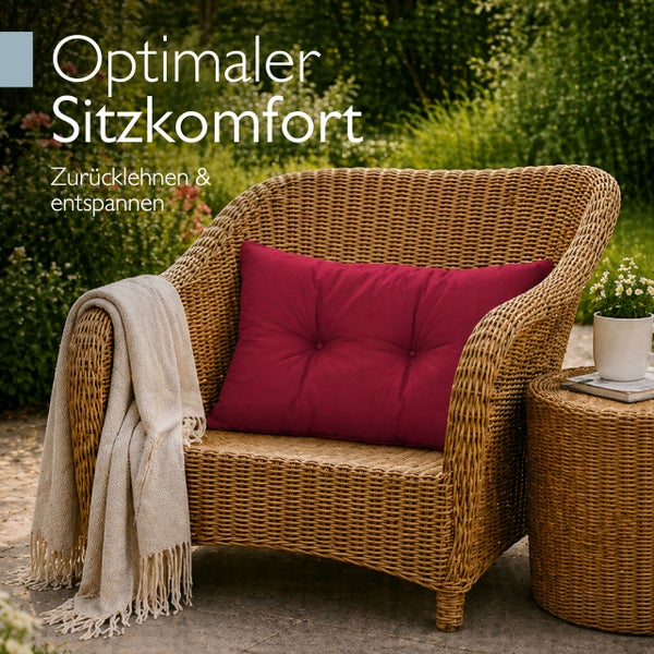Gartenstuhl aus Korbgeflecht mit einem Kissen und einer Decke auf einer Terrasse vor Gartenkulisse