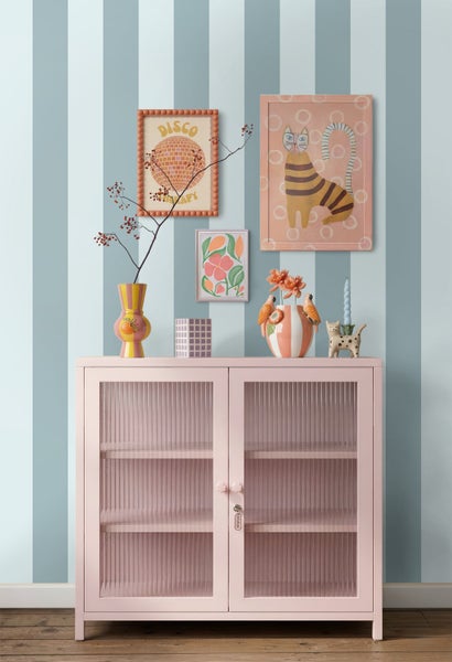 Organisiertes Zimmer mit einem rosa Schrank mit geriffelten Glastüren vor einer gestreiften Wand mit eklektischen Kunstwerken und Vasen.
