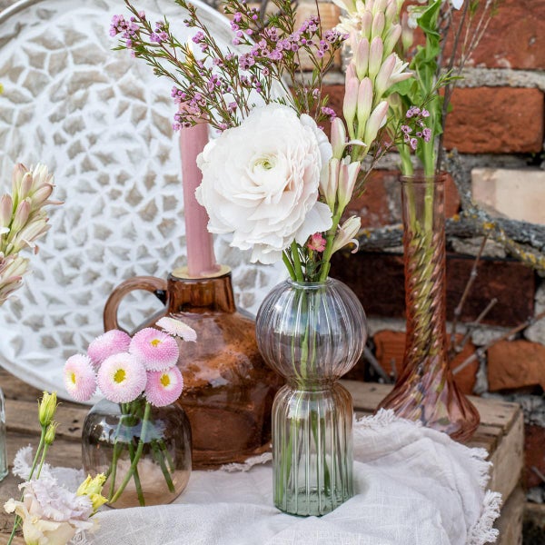 Dekorative Anordnung aus Glasvasen mit Ranunkeln, Gänseblümchen und Wachsblumen sowie einem Kerzenhalter auf einer Holzplatte vor einer Ziegelwand.