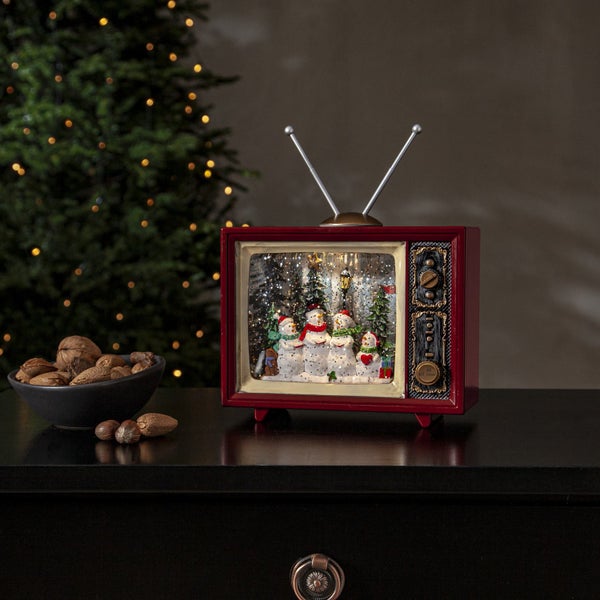 Dekorative Schneekugel in einem Fernsehgerät mit Schneemannszene, platziert auf einer Kommode. Eine Schale mit Nüssen steht daneben.