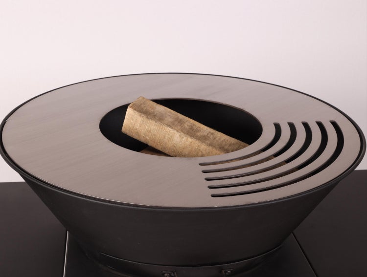 Runde Feuerschale mit Grillring aus Metall, Schlitzöffnungen für den Grill und einem Holzscheit in der Mitte.
