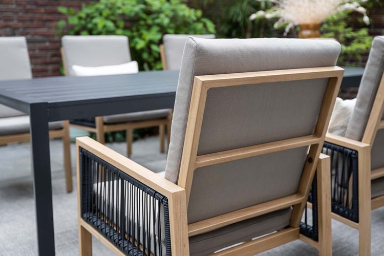 Gartenstuhl mit Holzrahmen, grauen Polstern und schwarzer Seilbespannung an den Armlehnen an einem grauen Gartentisch auf einer Terrasse.