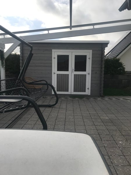 Graues Gartenhaus aus Holz mit einer weißen Doppeltür auf einer gepflasterten Terrasse.