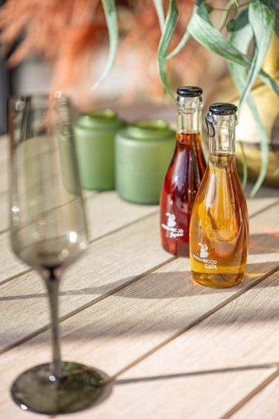 Sektglas auf einem Holztisch neben kleinen Flaschen Primasecco Lilly und grünen Windlichtern vor Pflanzen im Hintergrund.