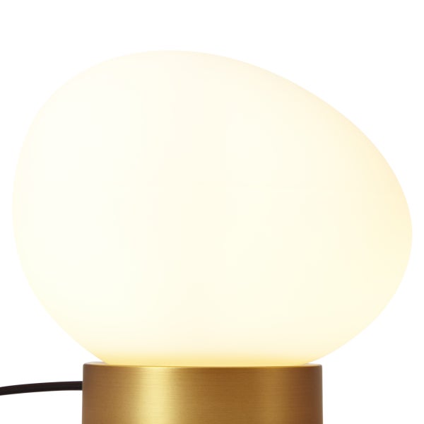 Tischleuchte mit ovalem Glasschirm in Kieselsteinform und zylindrischem Metallfuß in Messingoptik, warmweiß leuchtend.