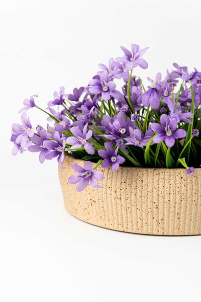 Künstliche violette Blumen in einer beigen strukturierten Schale mit vertikalen Rillen.