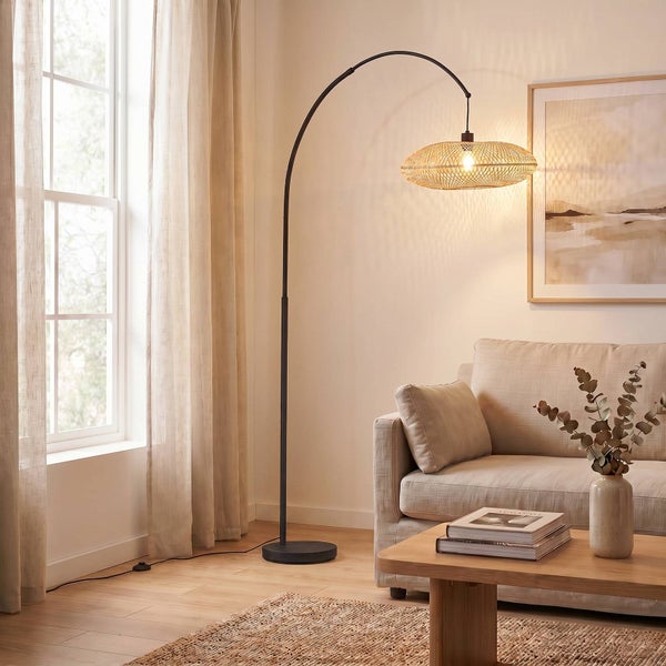 Bogenlampe mit schwarzem Metallgestell und Schirm aus Rattangeflecht in einem hellen Wohnzimmer mit Sofa und Holztisch.