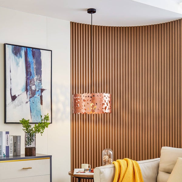 Moderner Wohnraum mit einer kupferfarbenen Pendelleuchte mit geometrischem Muster vor einer dekorativen Wand aus Holzlamellen.