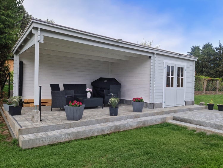 Modernes weißes Gartenhaus aus Holz mit überdachter Terrasse, Lounge-Set, Grill und Blumenkübeln auf einer Steinterrasse.