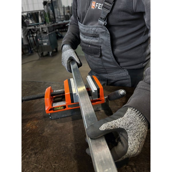 Eine Person in Arbeitskleidung mit FEIN Logo fixiert ein Metallprofil in einer orangefarbenen Klemmvorrichtung mit Wasserwaage auf einer Werkbank.