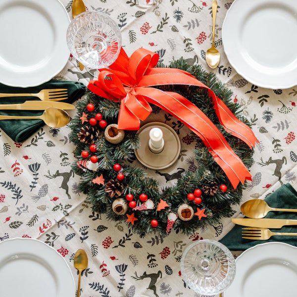 Festlich gedeckter Tisch mit Weihnachtskranz, roter Schleife, goldfarbenem Besteck, weißen Tellern und gemusterter Tischdecke aus der Vogelperspektive.