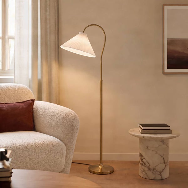 Moderne Stehlampe mit geschwungenem Messinggestell und plissiertem Lampenschirm im hellen Wohnzimmer neben einem Sessel und Marmorbeistelltisch.
