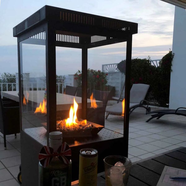 Moderner Außen-Gas-Kamin mit Glaswänden und brennender Flamme auf einer Terrasse mit Gartenmöbeln bei Dämmerung.