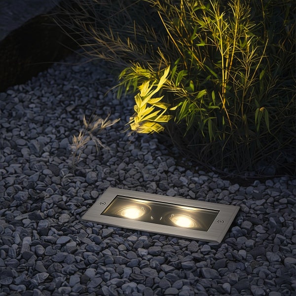Rechteckiger Bodeneinbaustrahler aus Metall mit zwei Lichtquellen, eingebettet in ein Kiesbeet zur Beleuchtung von Pflanzen im Garten.