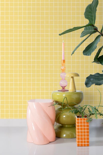 Dekoratives Stillleben mit Tisch, Hocker, Kerzenständer und Pflanzen vor gelber Mosaikwand.