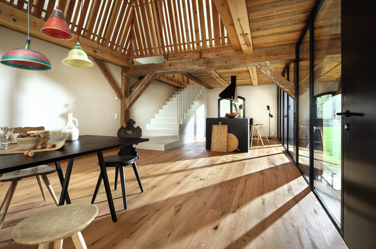 Heller, geräumiger Innenraum mit Eichenparkett, sichtbaren Holzbalken, einem schwarzen Esstisch mit bunten Pendelleuchten und einer modernen weißen Treppe.
