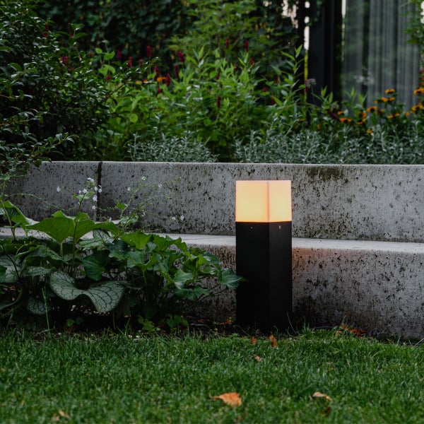 Schwarze LED-Wegeleuchte mit warmweißem Licht in einem Garten neben einer Betonmauer und Pflanzen.