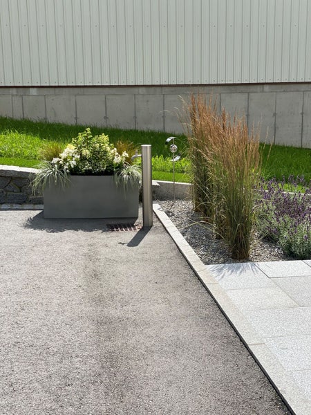 Moderne Garten-Wasserzapfsäule aus Edelstahl neben einem grauen rechteckigen Pflanzkasten mit weißen Blumen, hohen Ziergräsern und Lavendel.