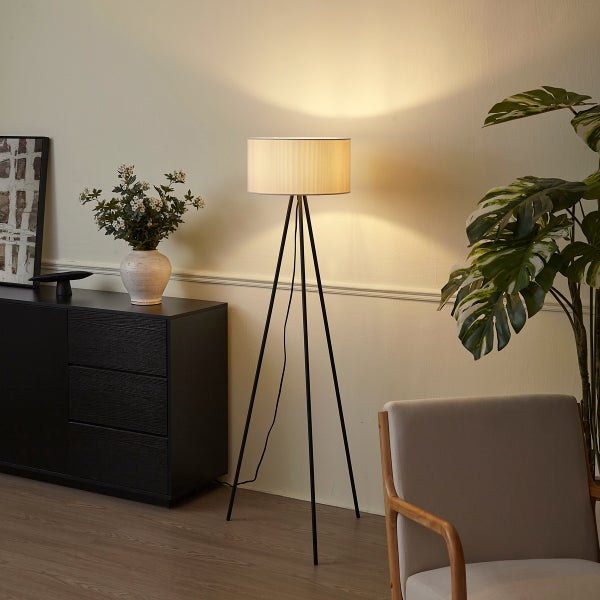 Moderne Dreibein-Stehleuchte mit Stoffschirm, beleuchtet, in einem Wohnzimmer mit schwarzer Kommode und Zimmerpflanze.