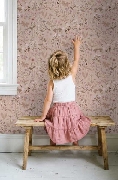 Vliestapete mit Wildblumen-Motiv im Kinderzimmer, ein Kind sitzt auf einer rustikalen Holzbank vor der tapezierten Wand.