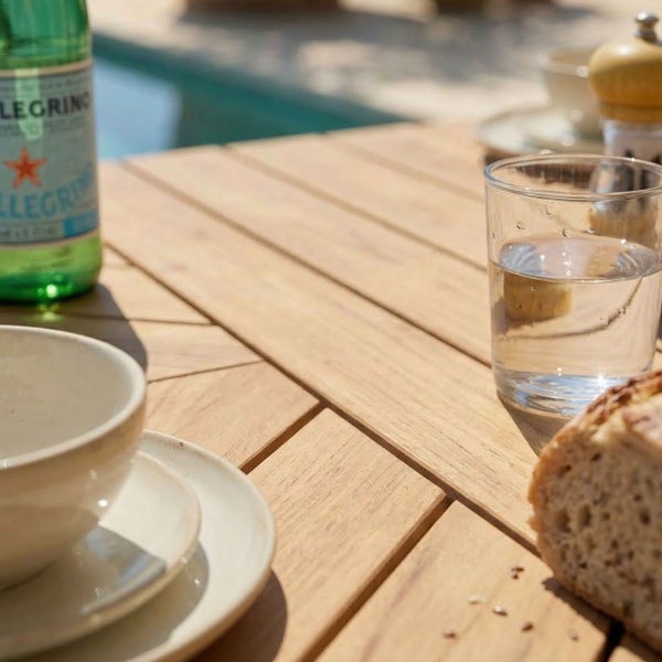 Detailansicht eines Gartentisches aus Holz mit Lattenstruktur, gedeckt mit Wasserglas, Schale und Brot im hellen Außenbereich.