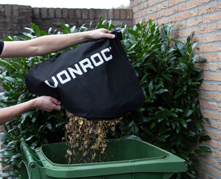 Eine Person leert einen Vonroc Laubsack mit Gartenabfällen in eine grüne Mülltonne vor einer Hecke.