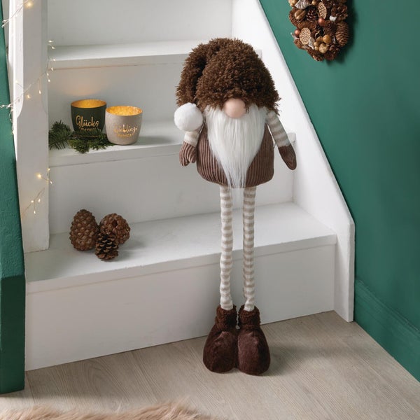 Dekorative Wichtelfigur mit langen Beinen und Mütze als Weihnachtsdekoration auf einer Treppe mit Tannenzapfen und Kerzen.
