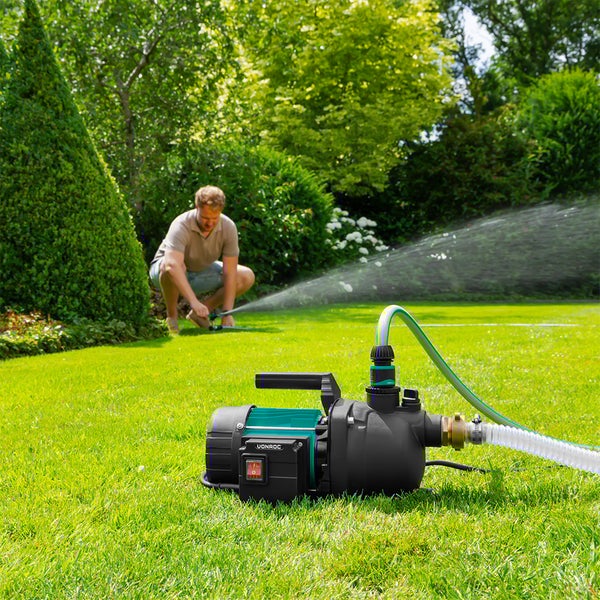 Vonroc Gartenpumpe auf einem Rasen mit angeschlossenen Schläuchen, im Hintergrund bewässert eine Person den Garten.