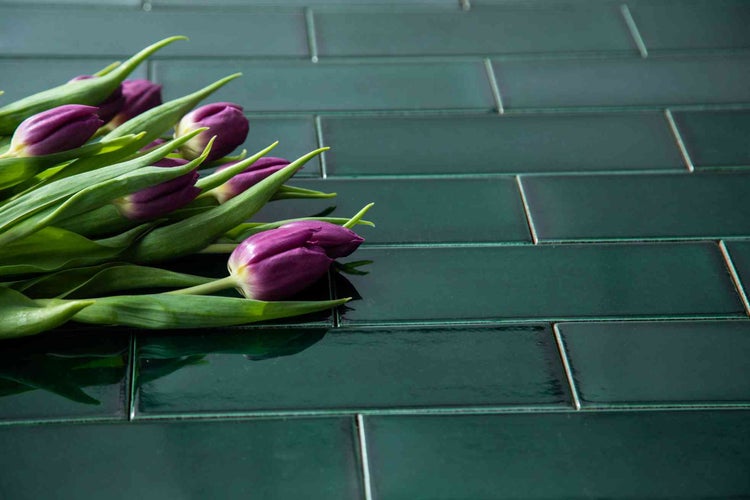 Rechteckige, dunkelgrüne, glänzende Wandfliesen aus Keramik im Mauerverband verlegt mit lila Tulpen.